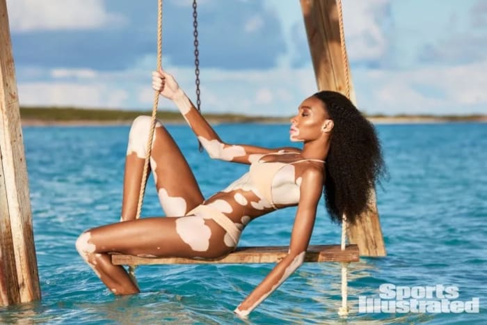 Winnie Harlow poses on a swing above the ocean, wearing a light tan two-piece bikini.